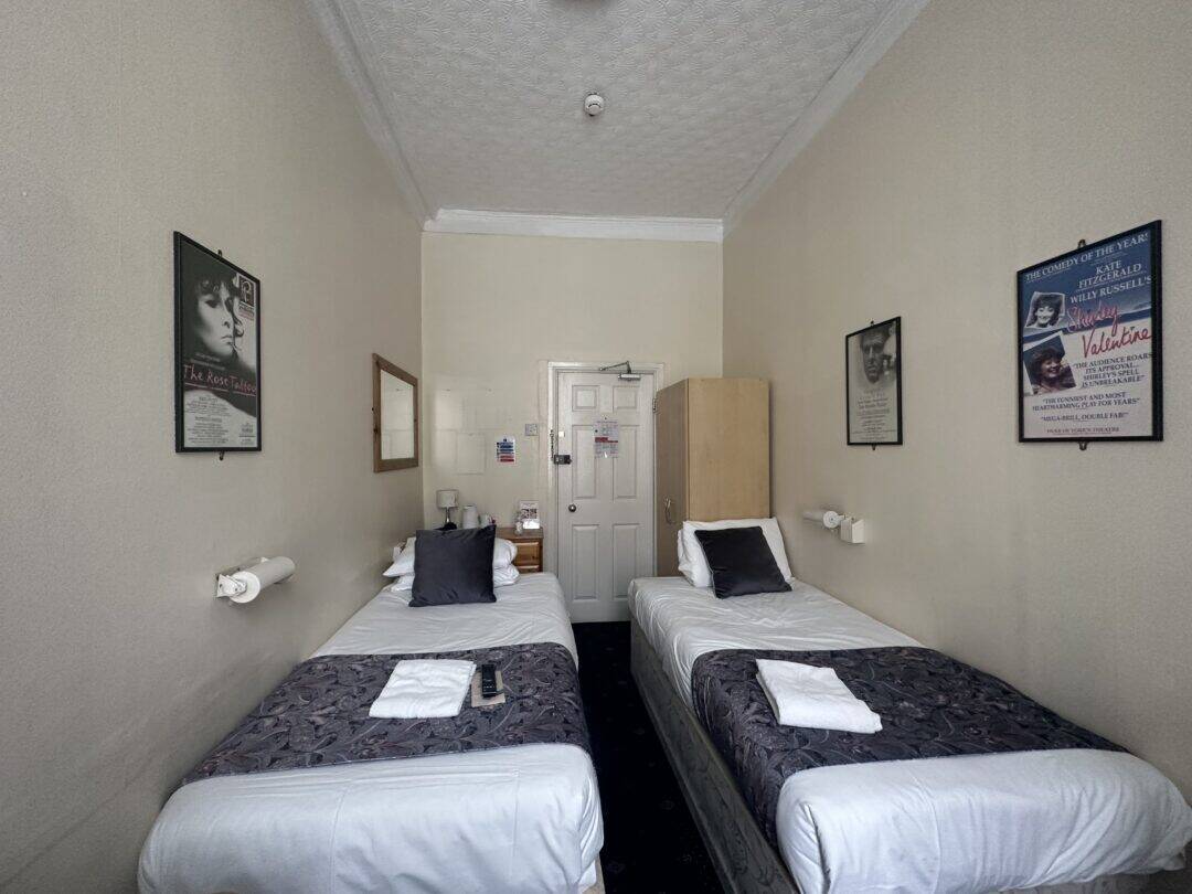 A small hotel room with two single beds, each with a folded towel and remote control on top. The room has beige walls, framed posters, a wardrobe, wall lamps, and a door at the far end.