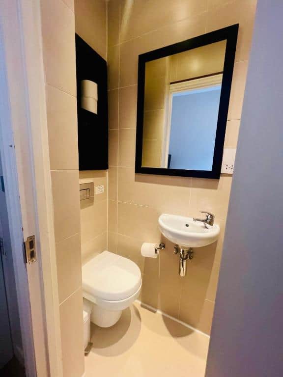 A small, modern bathroom with beige tiles, a wall-mounted toilet, a compact sink with a faucet, a toilet paper holder, and a large black-framed mirror above the sink. A black cabinet is mounted above the toilet.