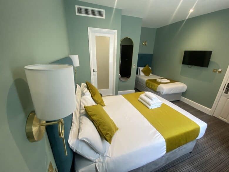 A modern hotel room with a double bed and a single bed, both with white linens and mustard-yellow accents. The room has green walls, a wall-mounted TV, a mirror, and neatly folded towels on the double bed.