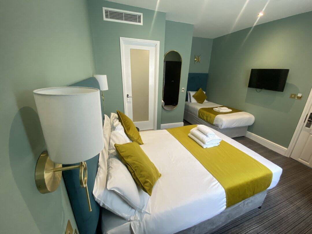A modern hotel room with a double bed and a single bed, both with white linens and mustard-yellow accents. The room has green walls, a wall-mounted TV, a mirror, and neatly folded towels on the double bed.