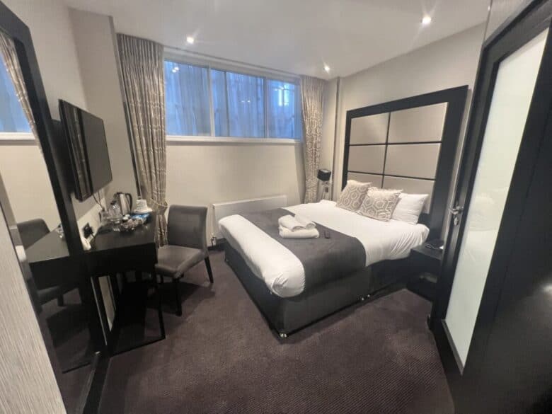 Modern hotel room with a double bed, two pillows, folded towels, a desk with a chair, wall-mounted TV, and large mirror. Beige curtains cover a window above the bed’s headboard. The decor is neutral and minimalistic.
