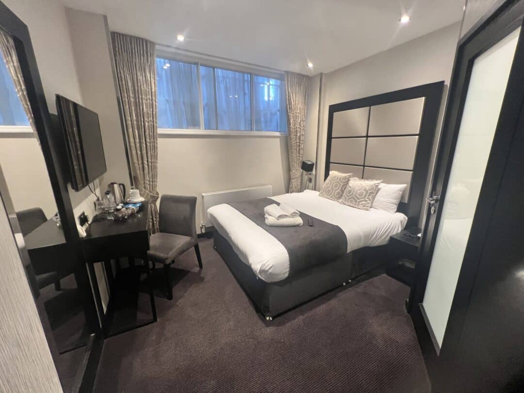 Modern hotel room with a double bed, two pillows, folded towels, a desk with a chair, wall-mounted TV, and large mirror. Beige curtains cover a window above the bed’s headboard. The decor is neutral and minimalistic.