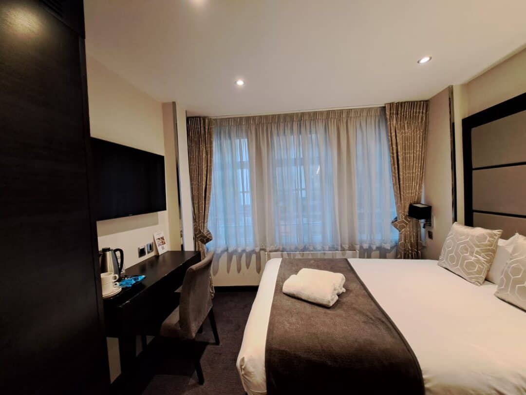 A modern hotel room with a neatly made double bed, brown and white bedding, a desk with a chair, a kettle, a mounted TV, and large windows with sheer curtains letting in daylight.