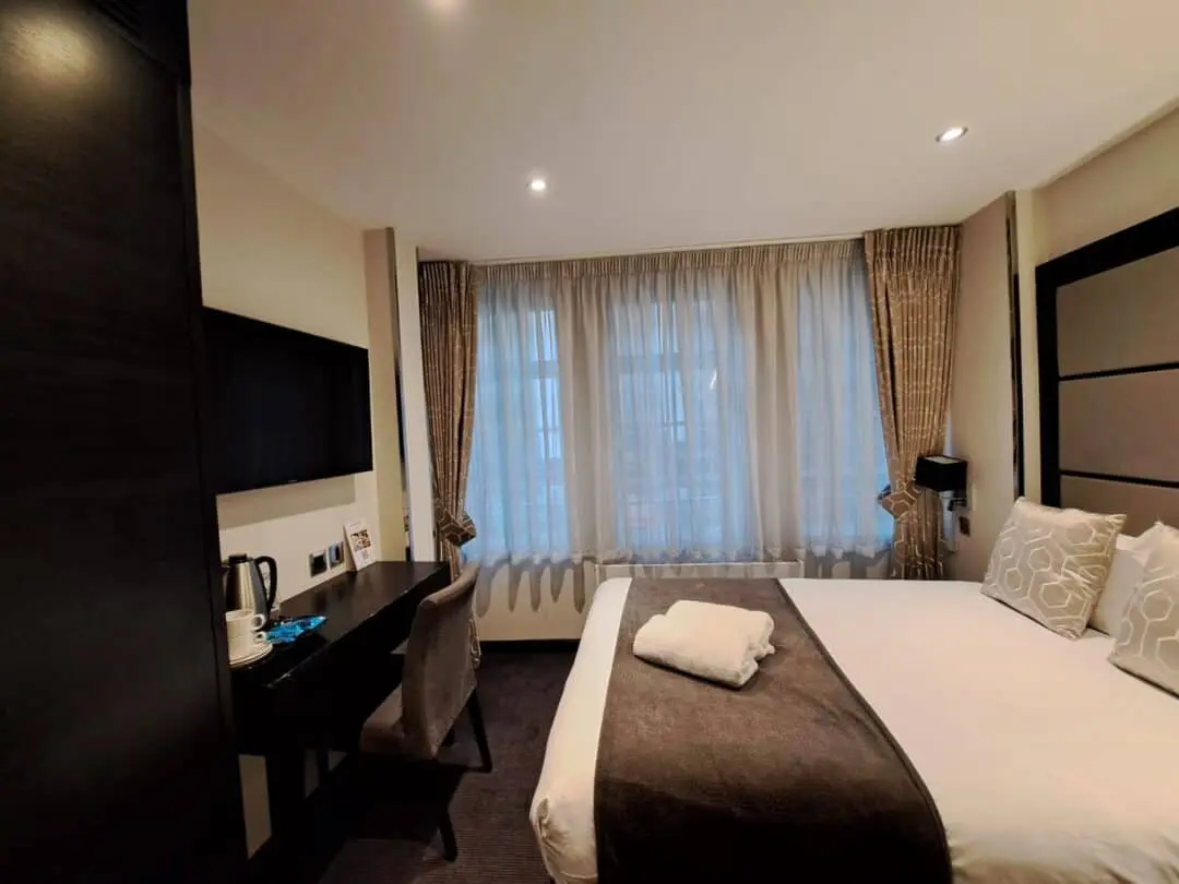 A modern hotel room with a neatly made double bed, brown and white bedding, a desk with a chair, a kettle, a mounted TV, and large windows with sheer curtains letting in daylight.