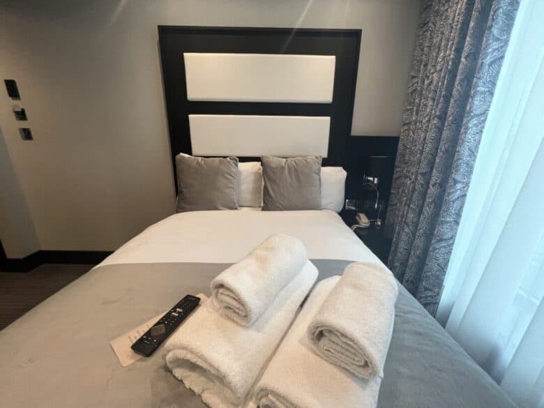 A neatly made bed with gray and white bedding, three pillows, and three rolled white towels on top. A TV remote rests on the bed, and light filters through patterned curtains by the window.