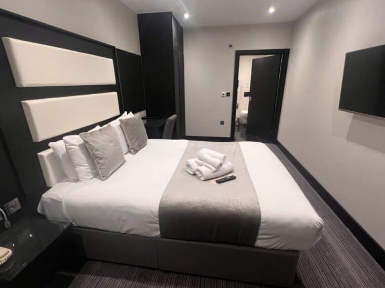 A modern hotel room with a double bed featuring white and gray bedding, several pillows, and towels neatly folded on top. A TV is mounted on the wall, and a door leads to another room.