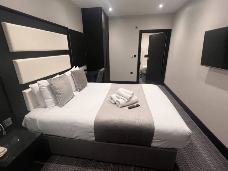 A modern hotel room with a double bed featuring white and gray bedding, several pillows, and towels neatly folded on top. A TV is mounted on the wall, and a door leads to another room.