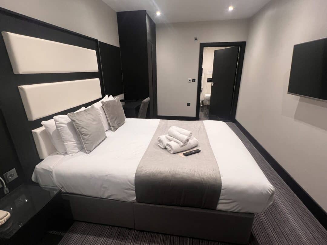 A modern hotel room with a double bed featuring white and gray bedding, several pillows, and towels neatly folded on top. A TV is mounted on the wall, and a door leads to another room.