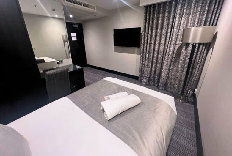 Modern hotel room with a neatly made bed, two rolled white towels on top, a wall-mounted TV, floor-length patterned curtains, a standing lamp, and a mirrored desk with a chair near the doorway.