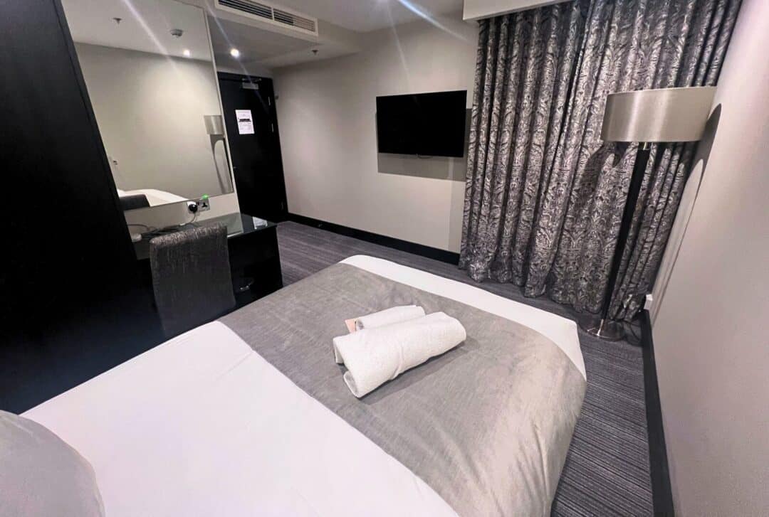 Modern hotel room with a neatly made bed, two rolled white towels on top, a wall-mounted TV, floor-length patterned curtains, a standing lamp, and a mirrored desk with a chair near the doorway.