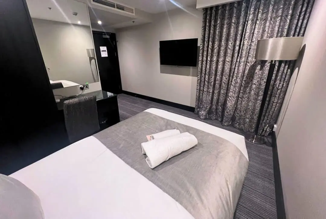 Modern hotel room with a neatly made bed, two rolled white towels on top, a wall-mounted TV, floor-length patterned curtains, a standing lamp, and a mirrored desk with a chair near the doorway.