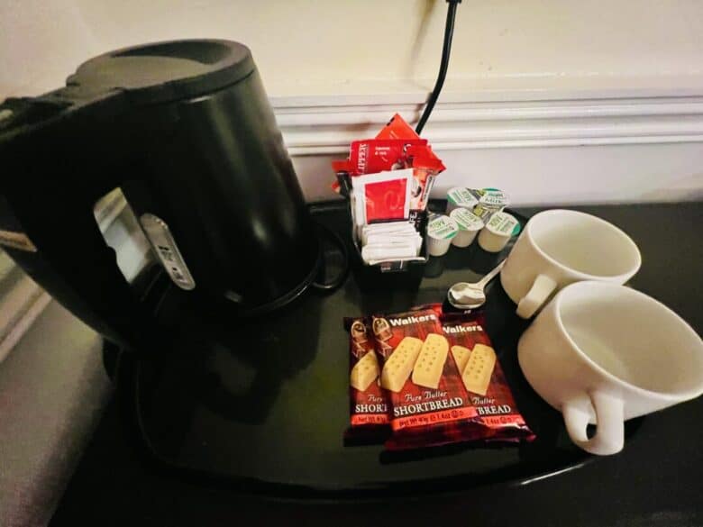 A black electric kettle, two white mugs, packets of Walkers shortbread, tea bags, sugar packets, and small coffee creamers are arranged on a black countertop.