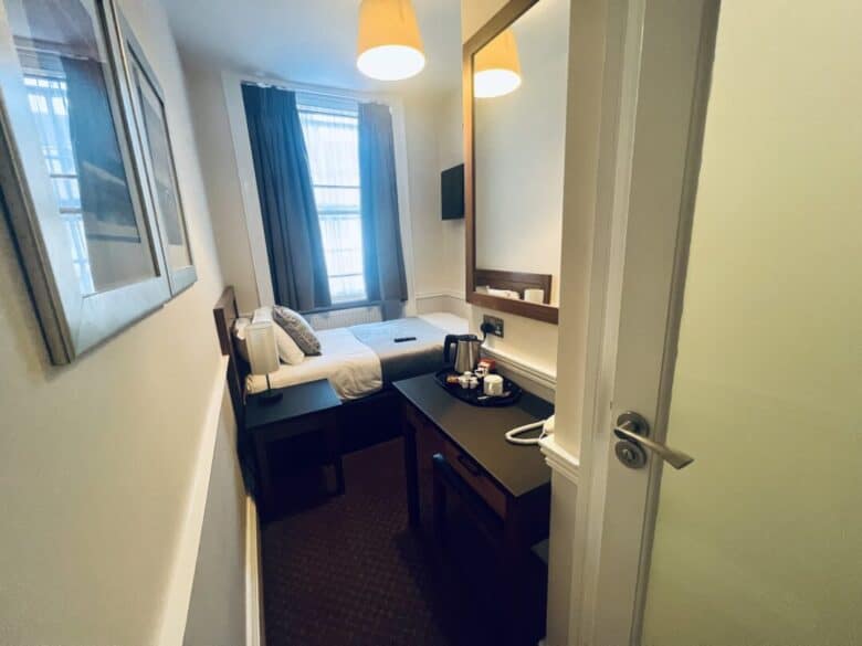 A small, tidy hotel room with a single bed by the window, a desk with a mirror, a tea kettle, mugs, and two pendant lights hanging from the ceiling. Curtains frame the window, and a painting hangs on the left wall.
