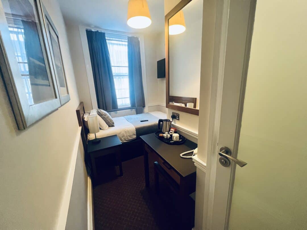A small, tidy hotel room with a single bed by the window, a desk with a mirror, a tea kettle, mugs, and two pendant lights hanging from the ceiling. Curtains frame the window, and a painting hangs on the left wall.