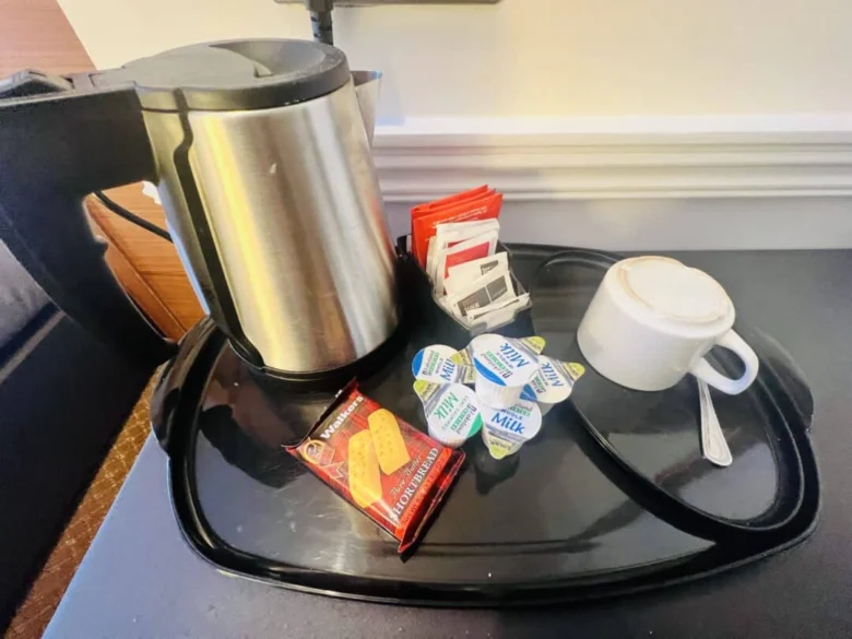 A metal electric kettle, packets of tea and coffee, creamers, a wrapped biscuit, a white cup with a saucer and spoon are arranged on a black tray on a countertop.