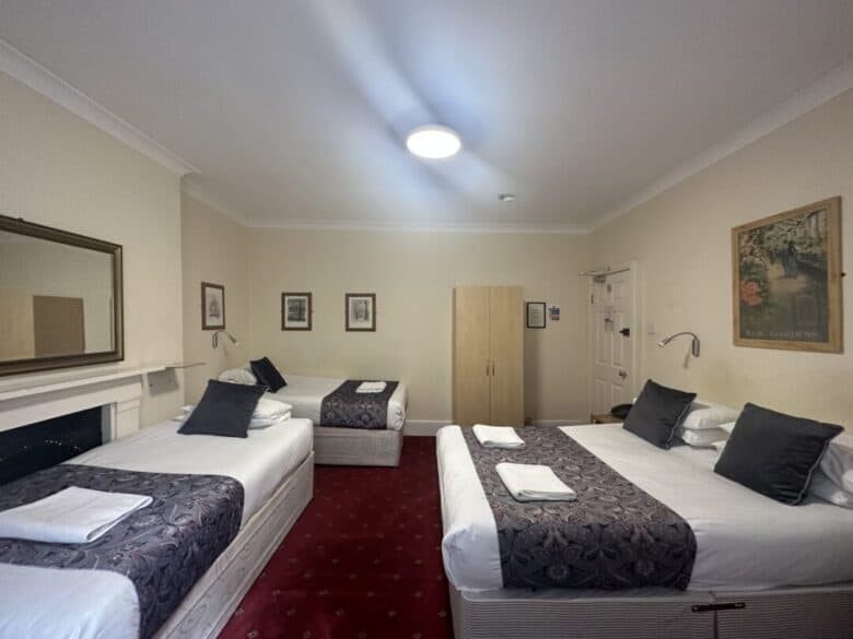 A hotel room with three beds—two doubles and one single—each neatly made with white linens, gray pillows, and patterned throws. The room has red carpet, framed art on beige walls, a wardrobe, and a large mirror.