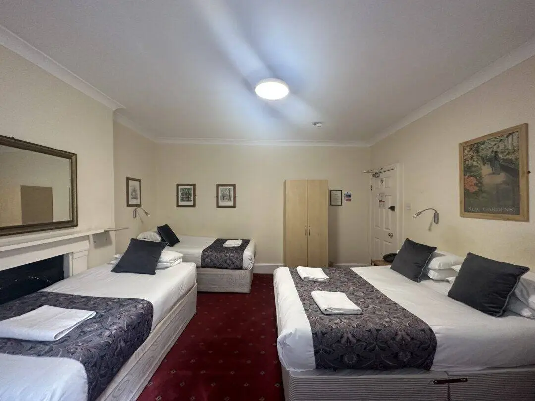 A hotel room with three beds—two doubles and one single—each neatly made with white linens, gray pillows, and patterned throws. The room has red carpet, framed art on beige walls, a wardrobe, and a large mirror.