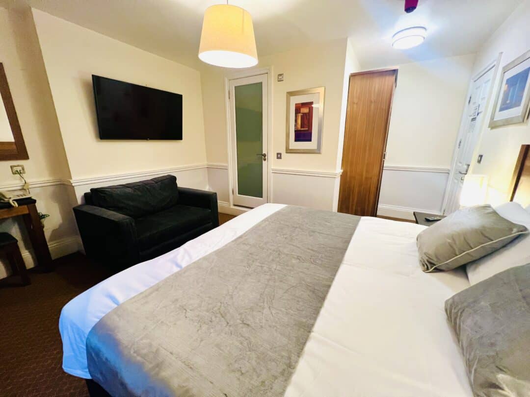 A cozy hotel room with a neatly made double bed, gray and white bedding, a dark sofa, wall-mounted TV, framed artwork, a wardrobe, and soft lighting from ceiling fixtures.
