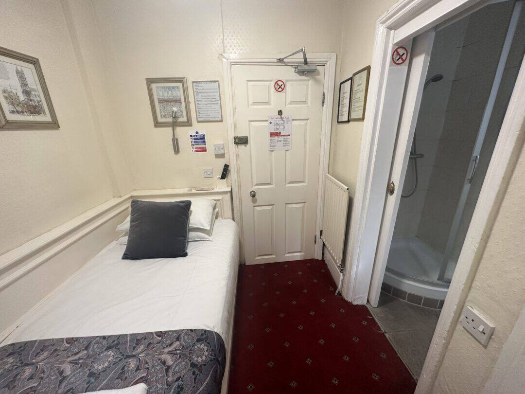Small hotel room with a single bed, two pillows, framed pictures on cream walls, a red carpet, and a door leading to a compact bathroom with a shower visible through an open doorway.