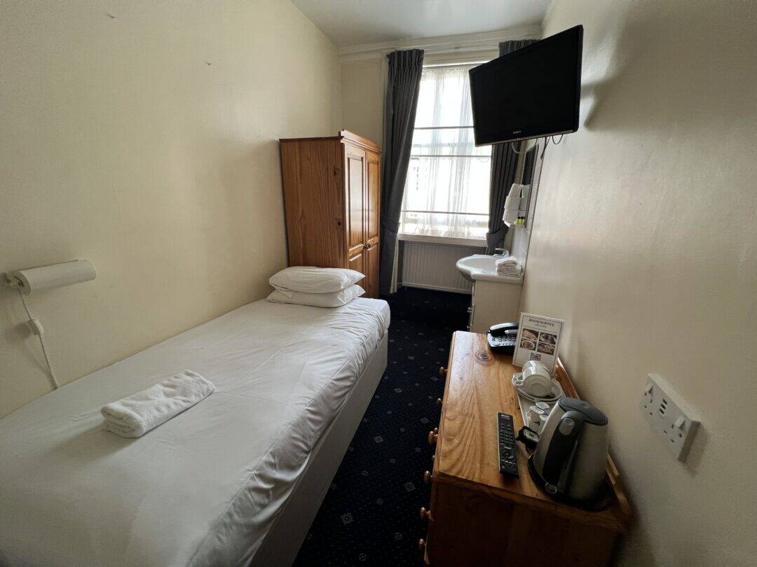 A small, simply furnished hotel room with a single bed, wooden wardrobe, desk with a kettle and tea set, wall-mounted TV, and a window with gray curtains letting in natural light.