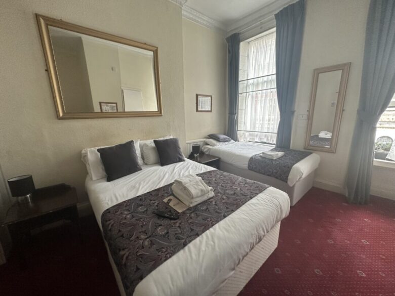 A hotel room with two beds, each with patterned runners and pillows, a bedside table with a lamp, a large window with curtains, a full-length mirror, and a wall mirror above one bed. Towels are folded on one bed.