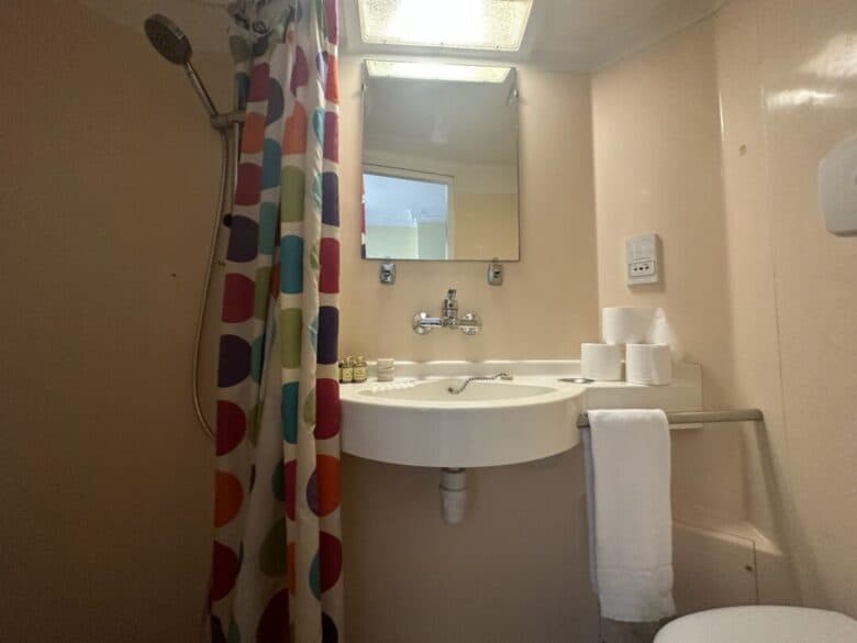 A small bathroom with a colorful shower curtain, wall-mounted showerhead, sink with mirror above, toilet paper rolls, toiletries, and a white towel hanging on a rail beneath the sink.