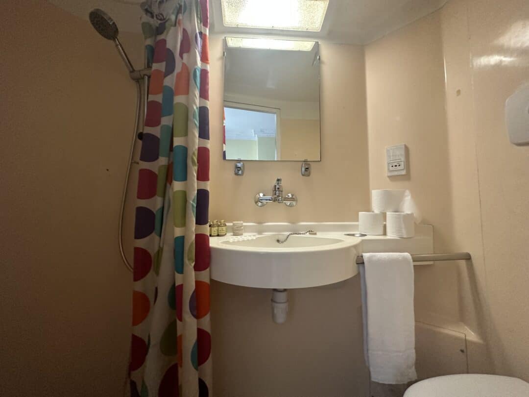 A small bathroom with a colorful shower curtain, wall-mounted showerhead, sink with mirror above, toilet paper rolls, toiletries, and a white towel hanging on a rail beneath the sink.