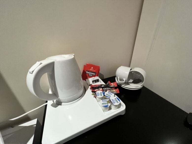 A white electric kettle sits on a tray with tea bags, sugar packets, and small milk containers. Nearby are stacked white cups with saucers and spoons on a black tabletop against a beige wall.