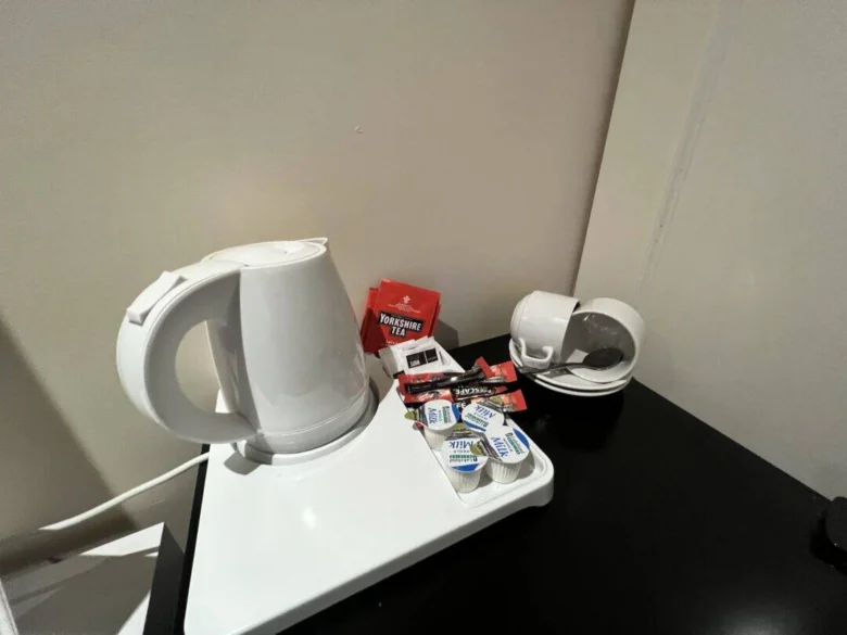 A white electric kettle sits on a tray with tea bags, sugar packets, and small milk containers. Nearby are stacked white cups with saucers and spoons on a black tabletop against a beige wall.