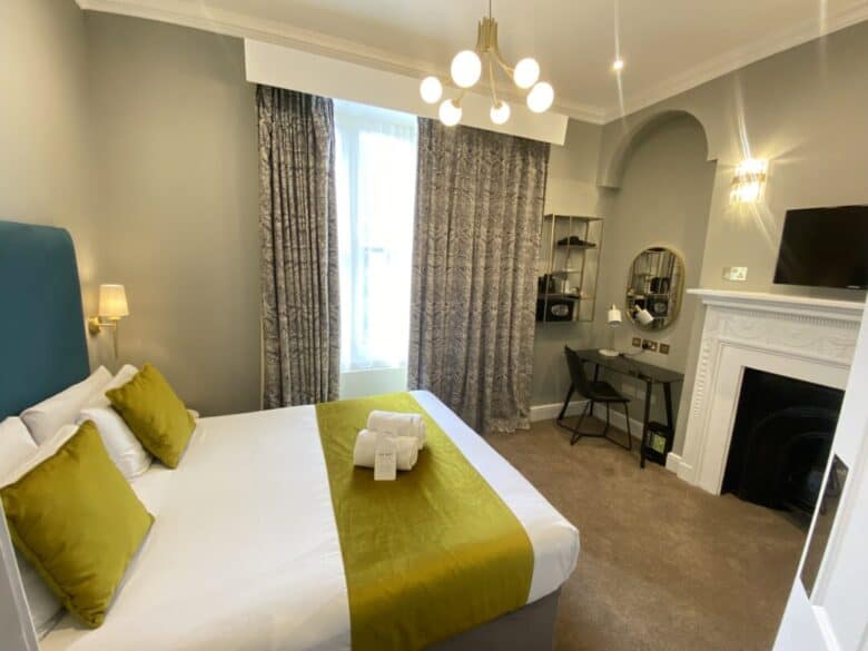 A modern hotel room with a double bed featuring green cushions and a matching throw, a wall-mounted TV, a desk with a round mirror, patterned curtains, and a fireplace under elegant lighting.
