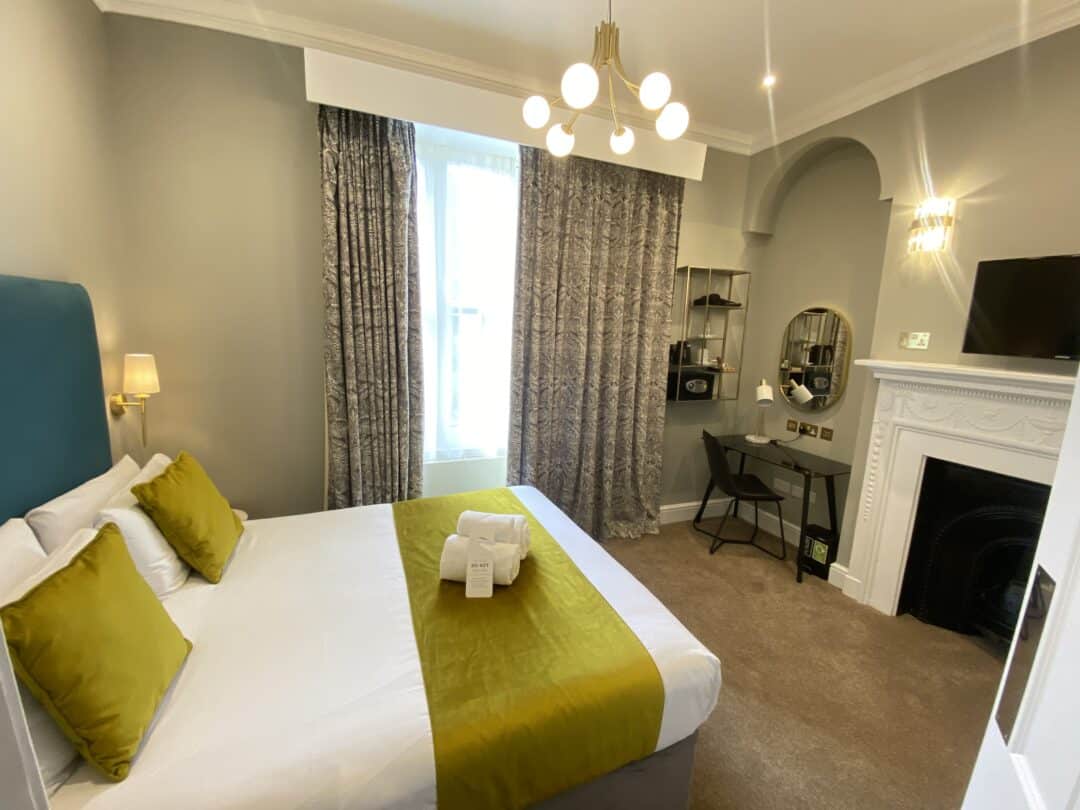 A modern hotel room with a double bed featuring green cushions and a matching throw, a wall-mounted TV, a desk with a round mirror, patterned curtains, and a fireplace under elegant lighting.