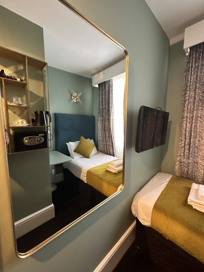 A small, modern hotel room with green walls, a neatly made single bed with yellow accents, a mounted TV, a large mirror, and shelves holding a kettle, cups, and a safe. Natural light enters through a window with patterned curtains.