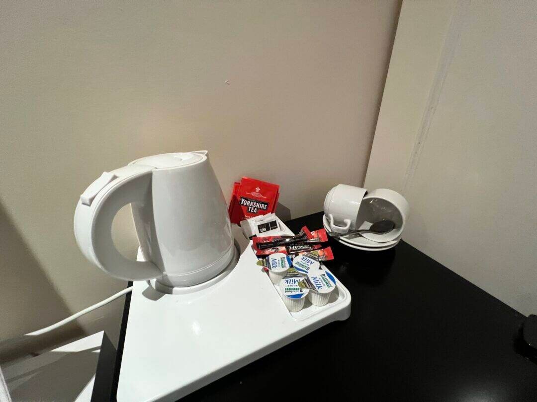 A white electric kettle sits on a tray with tea bags, sugar packets, and small milk containers. Nearby are stacked white cups with saucers and spoons on a black tabletop against a beige wall.