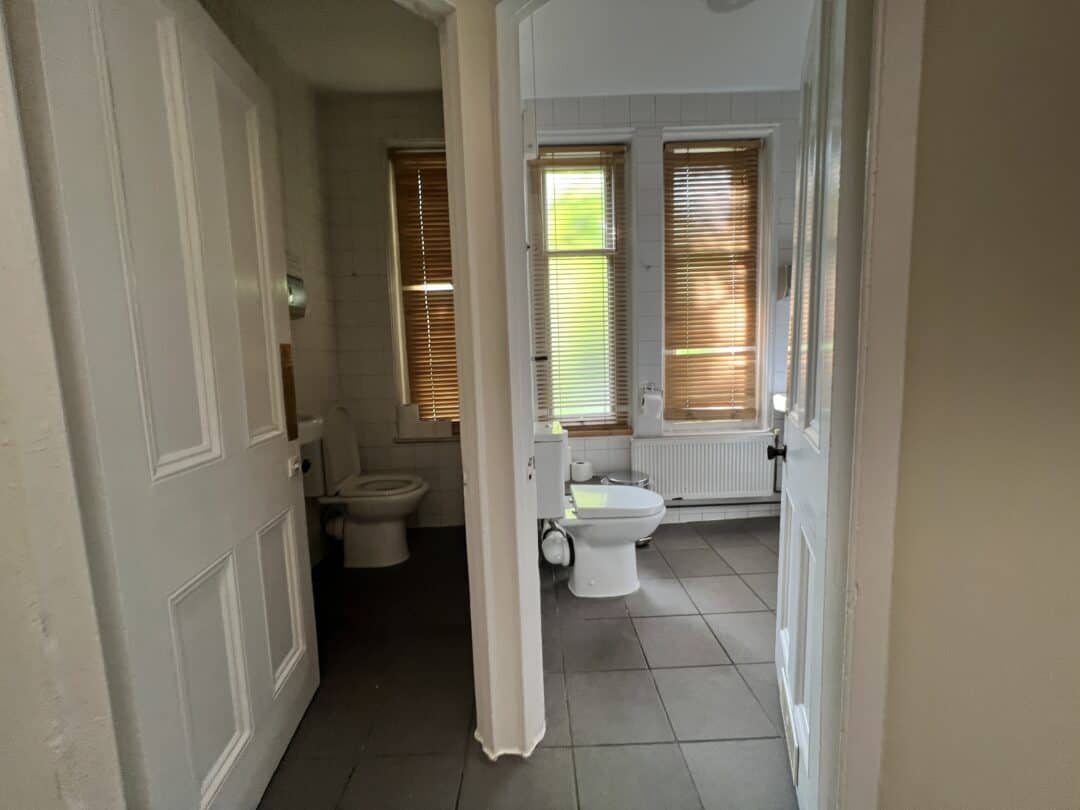 A view through open doors showing two adjacent bathrooms with tiled floors, toilets, sinks, and windows with wooden blinds letting in natural light.