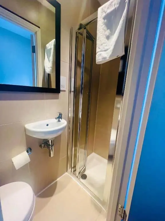 Compact bathroom with a small wall-mounted sink, toilet, and a glass shower enclosure. A towel hangs over the shower door, and a large mirror is above the sink. Beige tiles cover the walls and floor.