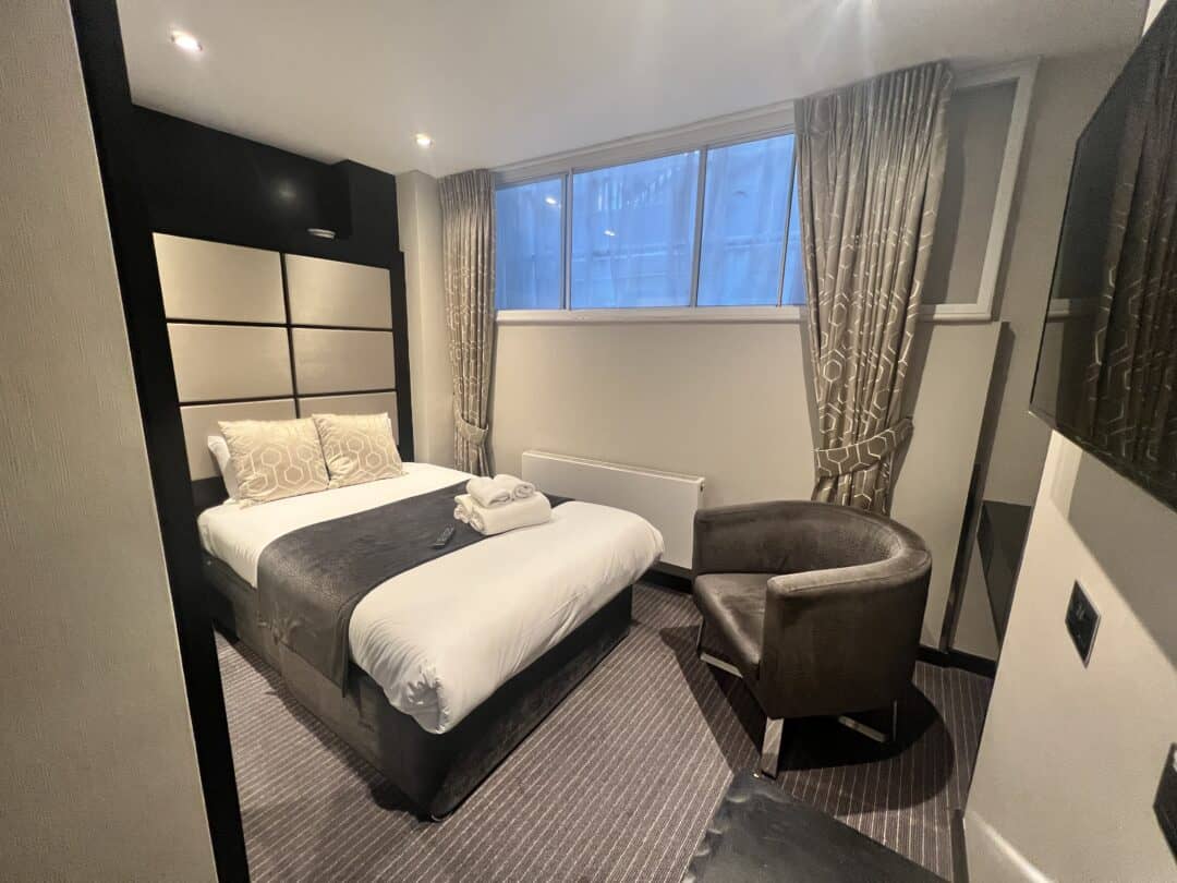 A neatly made hotel room with a double bed, two patterned pillows, a folded towel, brown curtains, a grey armchair, and a wall-mounted TV. Large window above the bed lets in natural light.