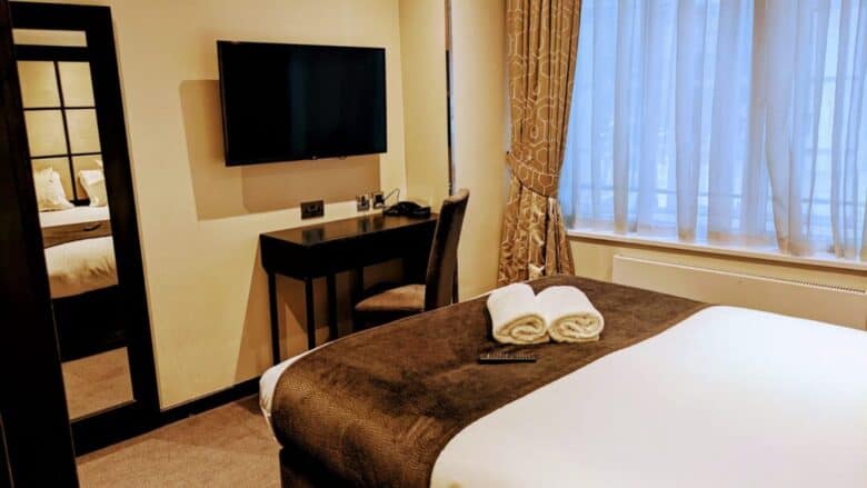 A modern hotel room with a neatly made bed, two rolled white towels, a TV mounted on the wall, a desk with a chair, and a large window with sheer curtains letting in daylight.