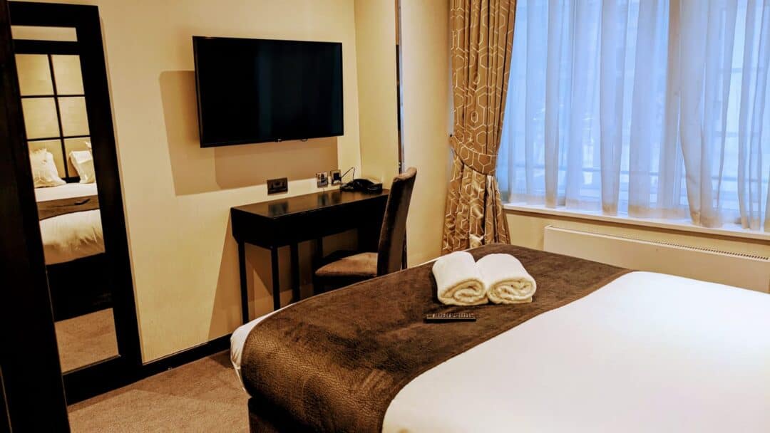 A modern hotel room with a neatly made bed, two rolled white towels, a TV mounted on the wall, a desk with a chair, and a large window with sheer curtains letting in daylight.