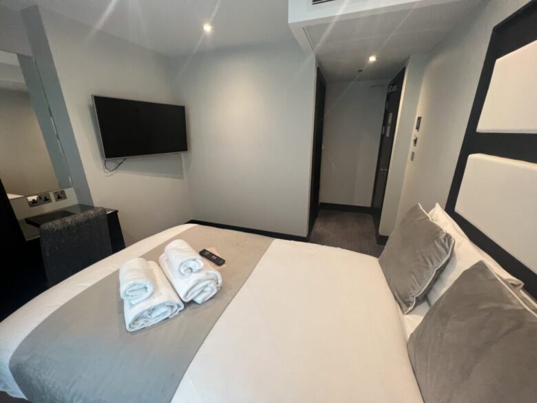A modern hotel room with a neatly made bed, grey and white bedding, three rolled towels, and a TV remote on the bed. A flat-screen TV is mounted on the wall, and the room has neutral grey décor.
