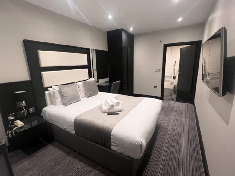 A modern hotel room with a neatly made double bed, folded towels on top, a wall-mounted TV, bedside tables, a desk, and an open door leading to a bright bathroom. The decor is neutral and minimalist.