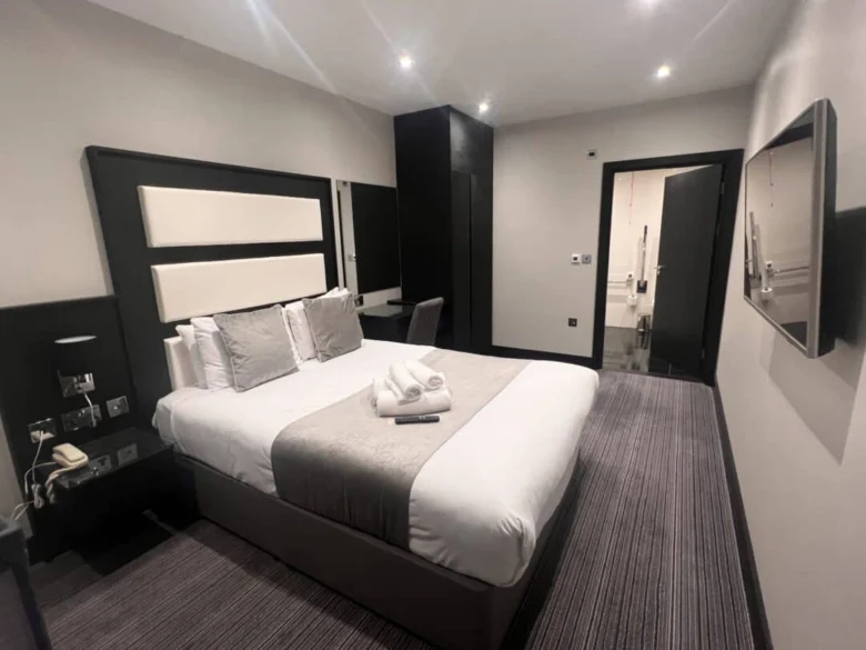 A modern hotel room with a neatly made double bed, folded towels on top, a wall-mounted TV, bedside tables, a desk, and an open door leading to a bright bathroom. The decor is neutral and minimalist.
