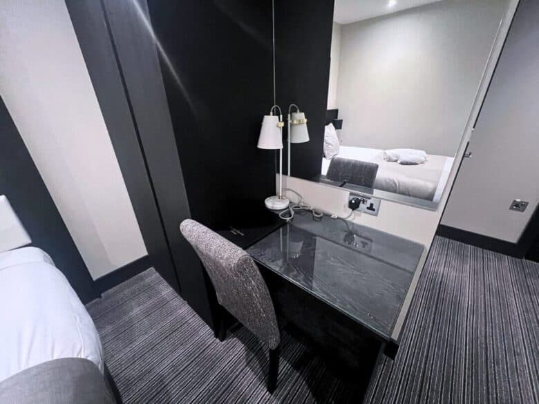 A hotel room featuring a bed with white linens, a black desk with a chair, a lamp, a mirror, and striped carpet flooring. The desk is positioned against a dark accent wall separating the sleeping area.