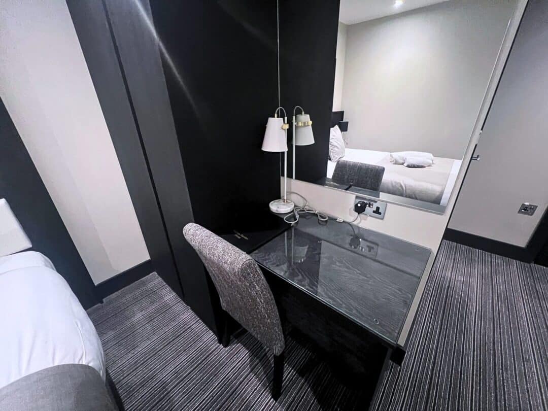 A hotel room featuring a bed with white linens, a black desk with a chair, a lamp, a mirror, and striped carpet flooring. The desk is positioned against a dark accent wall separating the sleeping area.