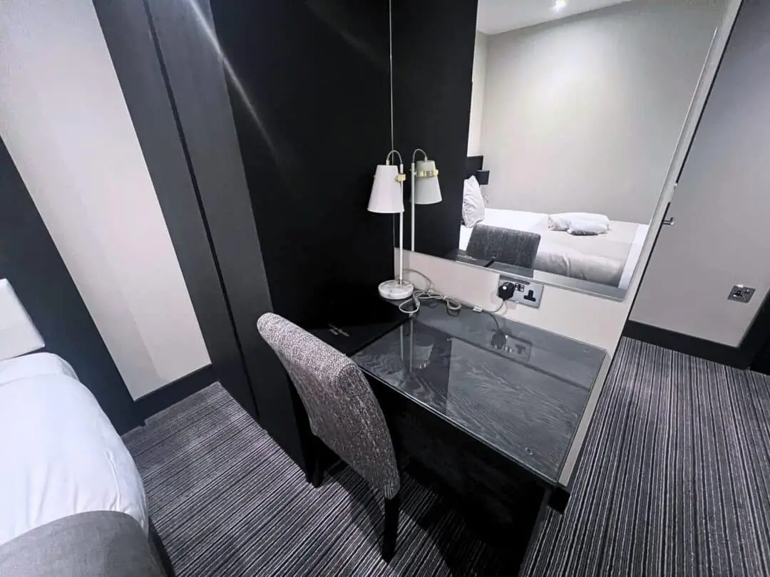 A hotel room featuring a bed with white linens, a black desk with a chair, a lamp, a mirror, and striped carpet flooring. The desk is positioned against a dark accent wall separating the sleeping area.