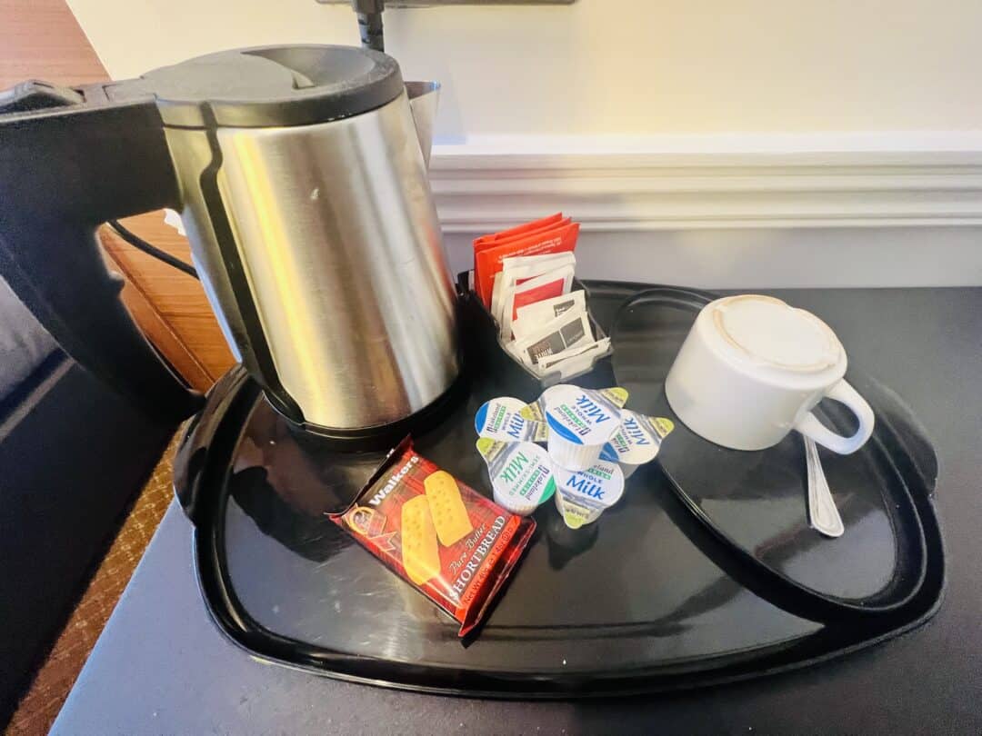 A hotel room beverage tray with an electric kettle, a cup and saucer with a spoon, tea and coffee sachets, small milk containers, and a pack of shortbread biscuits.