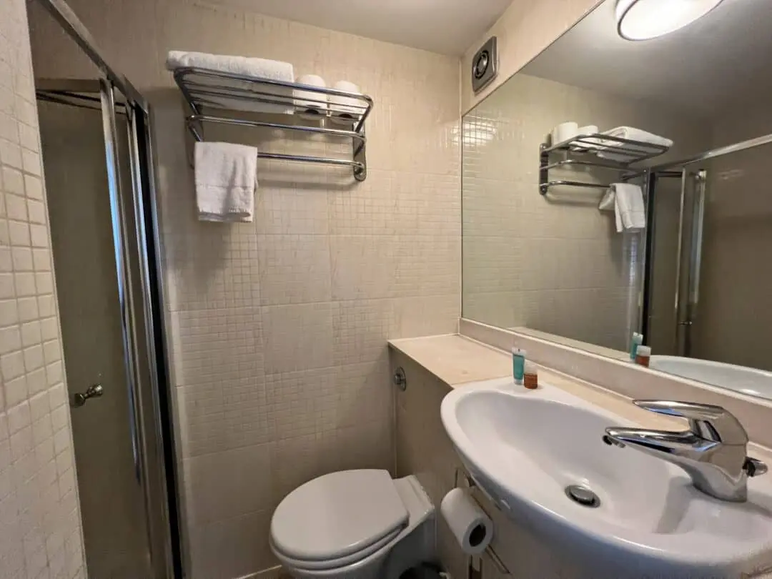 A small bathroom with beige tiled walls, a toilet, a wall-mounted sink with a mirror above it, a glass shower enclosure, and a metal shelf holding towels and toiletries.