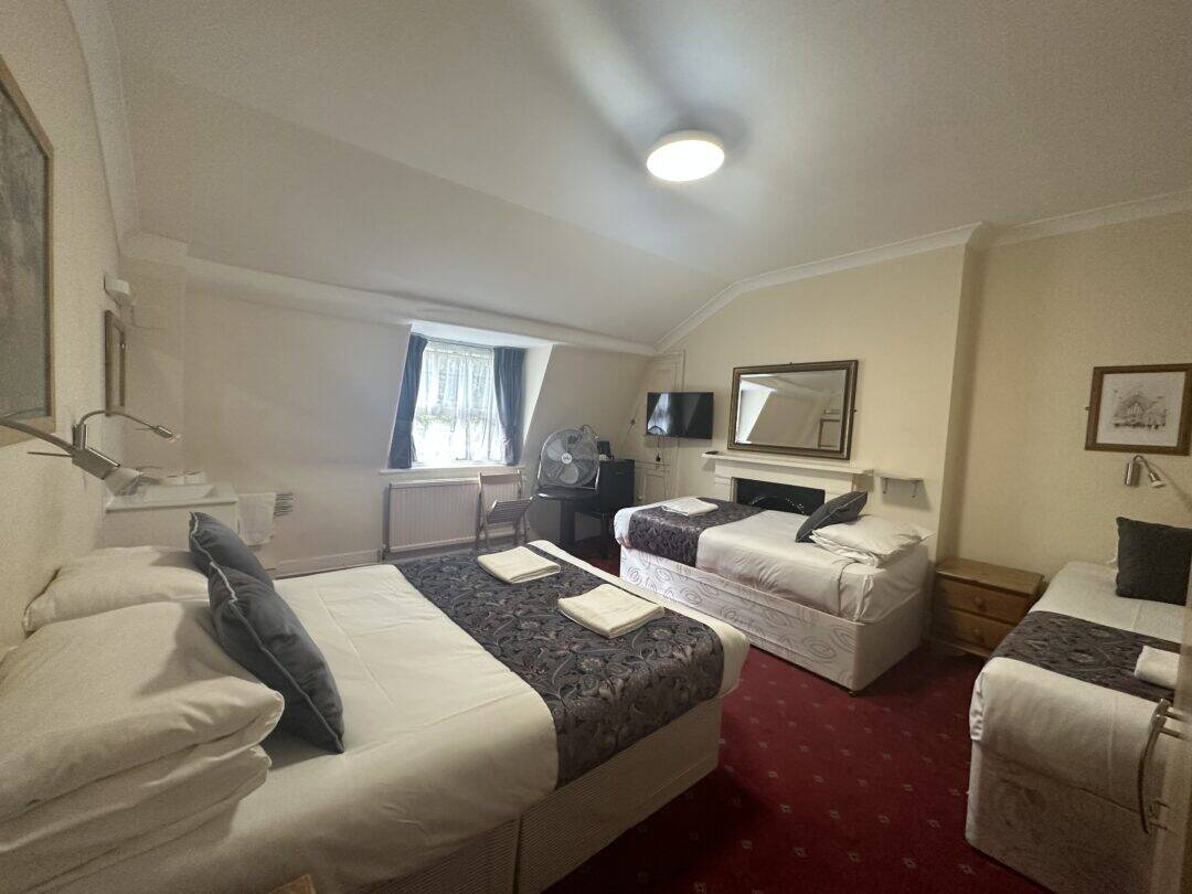 A cozy hotel room with two double beds and one single bed, each made with white linens and dark patterned throws. The room has red carpet, a window with sheer curtains, a mirror, TV, and a small table fan.