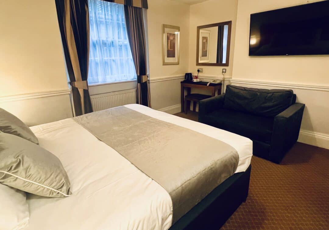 Cozy hotel room with a double bed, gray sofa, wall-mounted TV, small desk with chair, and a window with curtains. Neutral tones and soft lighting create a warm and inviting atmosphere.