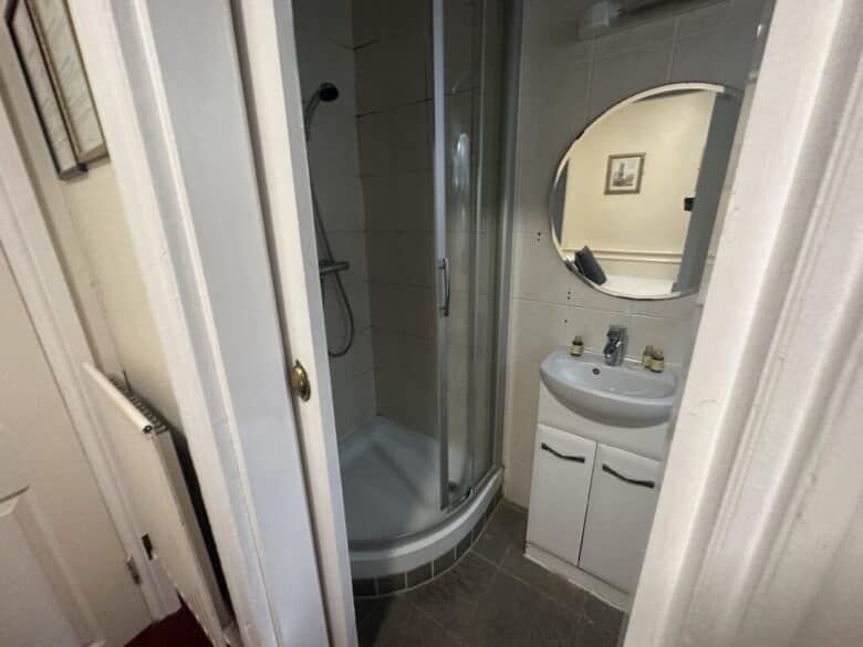 A small bathroom with a curved glass shower, a white sink cabinet, a round mirror above the sink, and bottles on the sink. A framed picture hangs on the wall, reflected in the mirror.