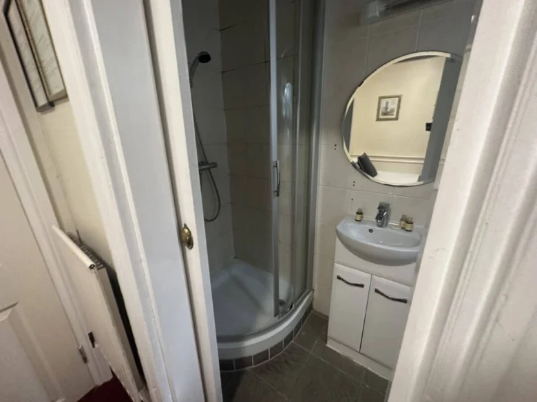 A small bathroom with a curved glass shower, a white sink cabinet, a round mirror above the sink, and bottles on the sink. A framed picture hangs on the wall, reflected in the mirror.